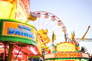 A fair with a ferris wheel, a corn dog stand, and a sign for cold drinks.