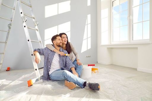 A smiling couple sitting on the floor near a ladder during a home renovation.